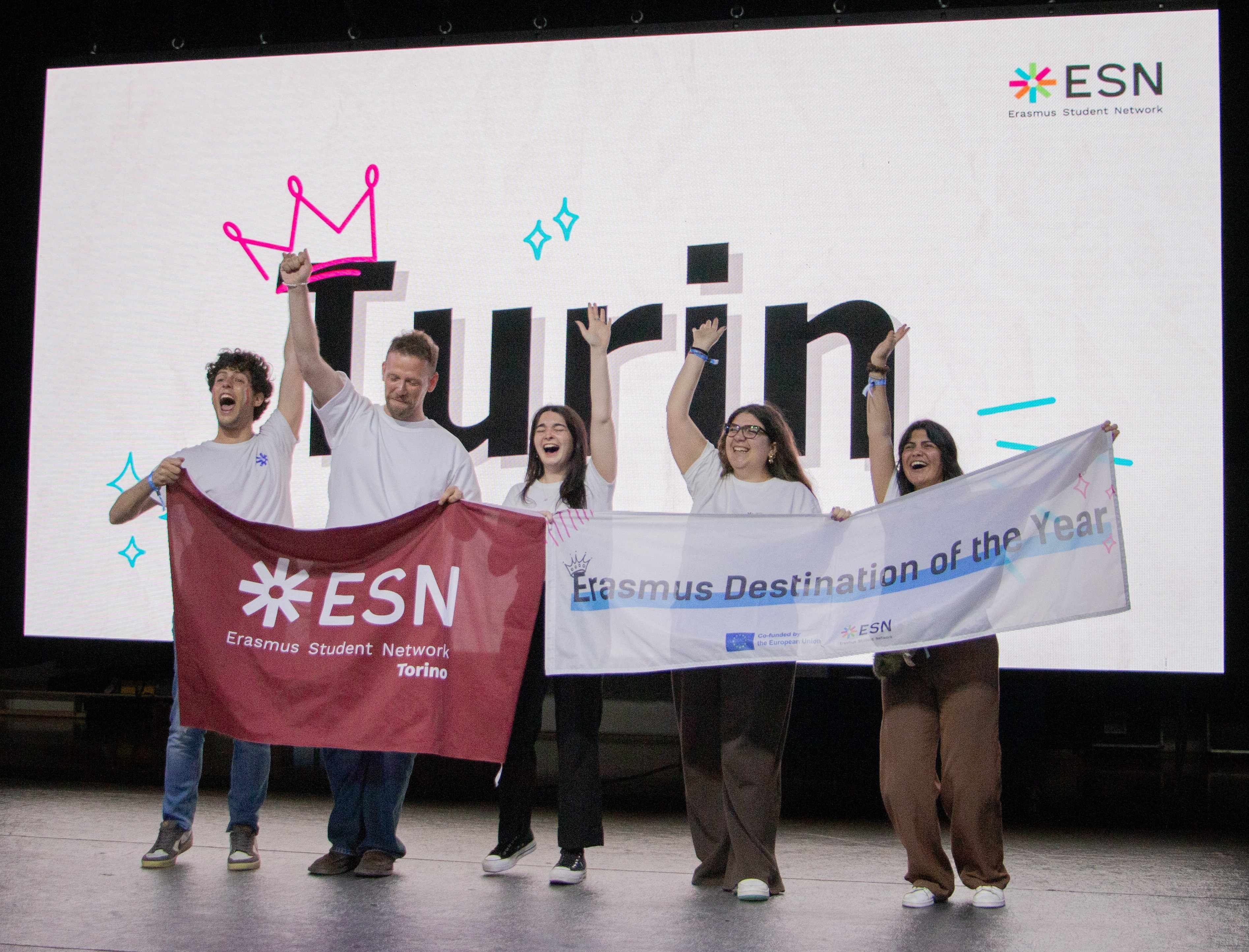 People cheering on a scenario in front of a big screen that says "Turin"